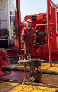 A man in red coveralls and a white helmet stands inferno of large machinery and next to a core barrel