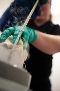 A green gloved hand is holding a fabric bag on a silver metal dipper. there is smoke around the dipper and the sample from the liquid nitrogen. the scientist's black t-shirt can be seen in the background.