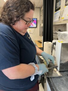 A scientist in a blue shirt and gloves uses a spatula to scrape the outside of a cylinder of core sample