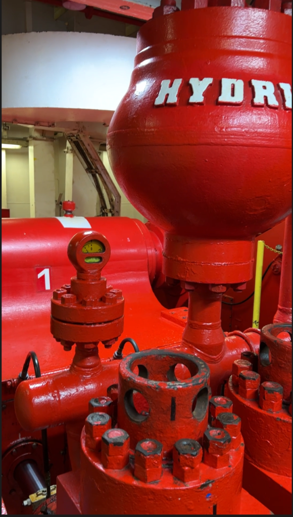 A arge red metal tank is in the top of the photo, it has "HYDRIL" in white lettering, A pipe from the bottom of this tank runs down into more pipes. They are all red metal.