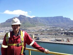 A man in red coveralls, a tan helmet, and a yellow harness stands infront of a yellow rail overlooking water and a mountain in the background.