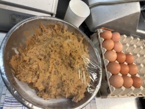 A bowl of dark cookie dough covered in clear plastic and a tray of brown eggs behind the bowl.