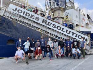 Eight people stand on a gangway decorated with a banner that says"JOIDES Resolution". a group of more people I standing on the pier infront of the gangway. All the people are wearing face masks.