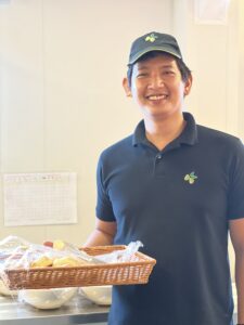 A smiling young man stands in a steward's uniform holding a basket of treats in the mess hall