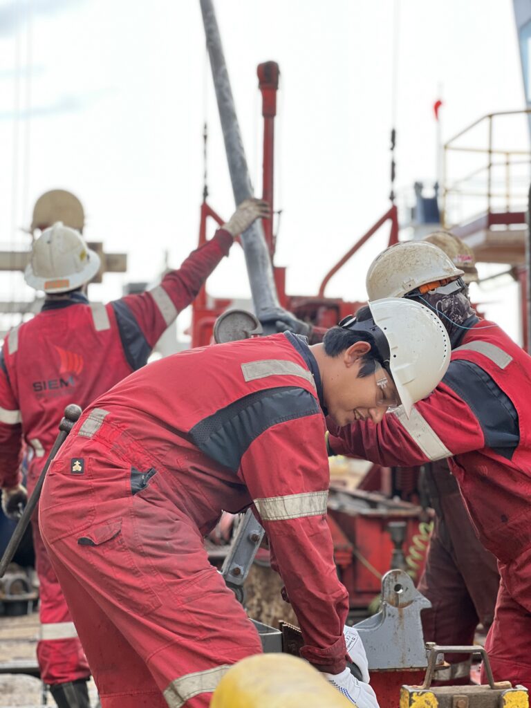 A smiling young man in red coveralls and a tan helmet stands bent over a core barrel on the drill floor. there are two en in red coveralls in the background holding the metal cylinder of the core barrel