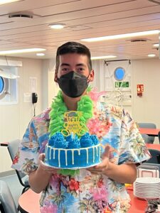 Young man with dark hair and a COVID mask wearing a Hawaiian shirt and lei and holding a blue frosted birthday cake.
