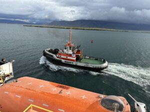 A tugboat in the harbor as taken from the deck of a ship. In the foreground is an orange lifeboat. The name of the tugboat is Magni and is shown on the side of the boat.