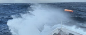 The bow of a ship with spray going over it. A wind sock is near