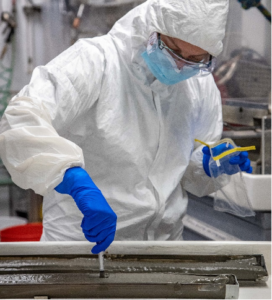 A person in a white body suit with a mask and goggles an blue plastic gloves is digging into sediment. 