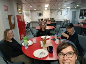 Four woman sitting around a table in the cafeteria of the galley of a ship. They are all smiling.