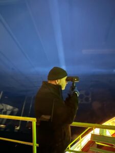 Man with a dark hat and cap, outside on the bridge of a ship looking through binoculars in the night.