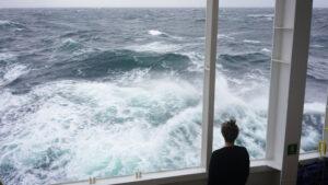 A man stands in the righthand foreground of the image looking off the side of the ship, waves fill the rest of the frame